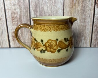 May include: A brown and cream ceramic creamer with a floral design. The creamer has a rounded body and a handle.