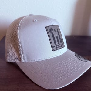May include: A light gray trucker hat with a mesh back and a curved brim. The front features a rectangular patch with a design and the text "PUERTO RICO BORIKEN". The hat is sitting on a wooden surface.
