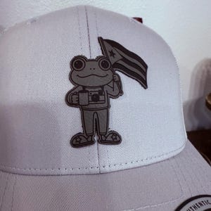 May include: A light gray baseball cap featuring a cartoon frog holding a flag and a camera. The frog is wearing star-shaped shoes. The cap has a curved brim and a snapback closure.