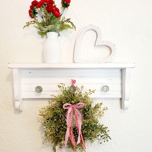 May include: A white, distressed wooden shelf with a small wreath, a vase of red flowers, and a heart-shaped decoration. The wreath has a pink and white checkered bow. The shelf has three decorative knobs.