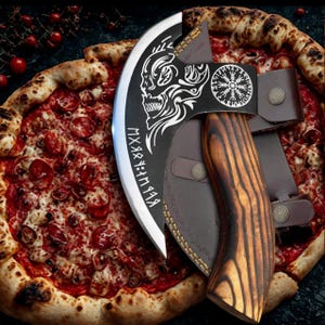 May include: A black and silver axe with a skull and runes design on the blade. The axe has a wooden handle with a brown leather sheath. The axe is resting on a pizza.