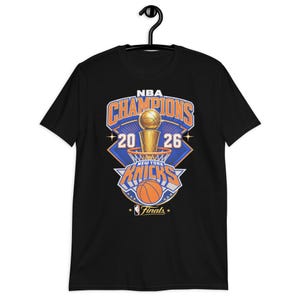 Peut inclure: Un t-shirt noir à manches courtes avec une impression graphique indiquant NBA CHAMPIONS 2026 NEW YORK KNICKS FINALS. Le motif comprend un trophée de basket doré, un panier de basket et un ballon dans des tons orange, bleu et blanc.