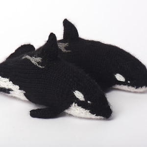 May include: Two handmade knitted orca whale plush toys. The whales are primarily black with white markings on their bellies and sides. They have small fins and a dorsal fin. The toys are lying on a white surface.