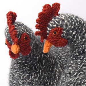 Op de afbeelding: Twee gebreide kippenfiguren. De kippen zijn grijs met witte vlekken en hebben rode kammen, oranje snavels en zwarte ogen. De kippen zijn gemaakt van een zacht, getextureerd garen. De achtergrond is wit.