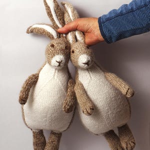 May include: Two hand-knitted stuffed rabbits featuring light brown and cream-colored yarn. Each rabbit has long ears with white inner sections, a cream-colored chest and belly, brown limbs, and embroidered facial features including eyes and a nose.