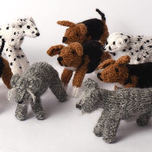 Könnte beinhalten: Eine Sammlung handgestrickter Hundefiguren in verschiedenen Rassen und Farben. Die Hunde umfassen einen Dalmatiner mit schwarzen Flecken, braune und schwarze Beagles und graue Schnauzer. Jeder Hund ist mit detaillierten Merkmalen gefertigt.