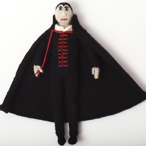 May include: A handmade knitted doll of a vampire, featuring a black cape and suit with red accents. The doll has a pale face, black hair, and a white collar. The cape is spread wide, and the doll has small black feet.