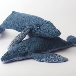 Puede incluir: Dos peluches de ballena azul tejidos. Las ballenas son en tonos de azul, con detalles texturizados y aletas estampadas. Una ballena está colocada encima de la otra, creando una disposición lúdica.