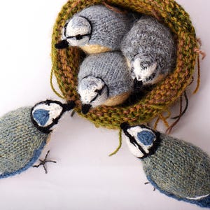 Op de afbeelding: Handgebreide blauwe gaaien met witte en zwarte accenten, rondom een nest met drie blauwe gaaijongen. Het nest is gemaakt van bruin, groen en geel garen. De vogels hebben blauwe vleugels en staarten.