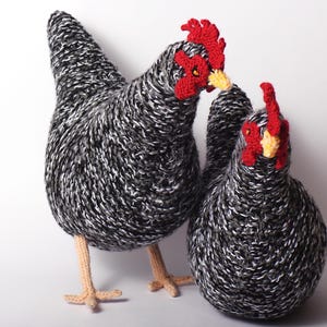 Könnte beinhalten: Zwei handgestrickte Hühner mit einem Körper aus schwarz-weiß meliertem Garn, einem leuchtend roten gehäkelten Kamm und Kehllappen sowie einem kleinen gelben Schnabel. Das größere Huhn steht auf zwei hellbeigen Strickbeinen mit Füßen.