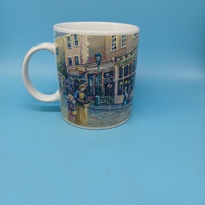 May include: White ceramic coffee mug featuring a detailed illustration of a street scene with buildings, people, and a Starbucks Coffee shop. The mug has a curved handle and a glossy finish. The scene is rendered in soft, muted colors.