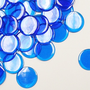 May include: A pile of blue glass game chips, also known as bingo chips or counters, on a white background. One chip is separated from the pile.