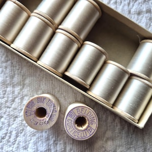 Puede incluir: Una colección de carretes de hilo de seda vintage en una caja de cartón. Los carretes son de color beige claro, con lados de madera y un brillo metálico. Se ven dos carretes individuales, con el texto "PURE SILK THREAD" y "BELDING CORTICELLI".