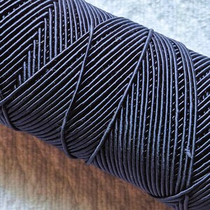 May include: Close-up of a spool of navy blue elastic cord. The cord is tightly wound, revealing a textured surface with diagonal and parallel lines. The cord appears to be made of a flexible material, suitable for various crafting projects.