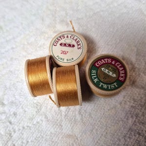 May include: Four spools of gold thread and two spools of Coats & Clark's thread. The thread spools are labelled "Pure Silk" and "Silk Twist". The thread is wound around wooden spools.