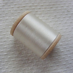May include: A spool of off-white sewing thread featuring a smooth, lustrous finish wrapped around a light-colored wooden cylinder. The thread is tightly wound, displaying a consistent, reflective texture across the entire surface of the spool.