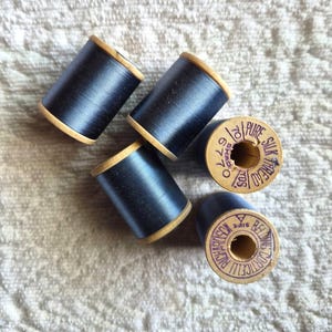 May include: Five spools of black thread with wooden sides. The spools are arranged on a textured, off-white surface. The ends of the spools have stamped text that reads "Pure Silk Thread" and "Belding Corticelli".