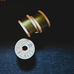 May include: Three spools of thread, two stacked and one separate, against a dark background. The thread is a muted olive green, with wooden spools. One spool has the text "BUTTON & BUTTONHOLE TWIST" and "SHADE 955".