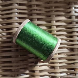 May include: A spool of bright green thread rests on a woven wicker surface. The thread is tightly wound around a wooden spool, showcasing a vibrant, emerald color. The background features a natural, light brown wicker pattern.