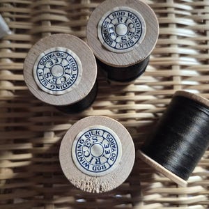 May include: Four spools of black silk thread with wooden bases. The spools are labeled with the text "Rod Winding Silk" and "50 Yards". The spools are arranged on a woven wicker surface.