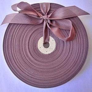May include: A spool of mauve ribbon with a matching bow. The ribbon is tightly wound and secured with a bow at the top. The ribbon is made of a woven material and is suitable for crafts and gift wrapping.