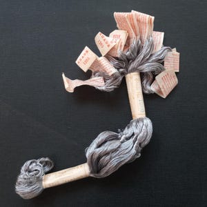 May include: A spool of silver gray embroidery floss with multiple paper labels that say "Shade 1200" and "DMC No. 1200".