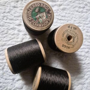 May include: Four spools of black Champion Silk thread by Gudebrod Bros. The wooden spools have printed labels with text and a shield logo. One spool reads "44 YDS" and another reads "Champion Silk".