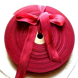 May include: A spool of deep red ribbon wound around a cardboard core. The ribbon is tied in a bow at the top of the spool.