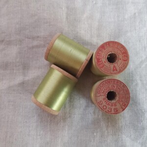 Puede incluir: Cuatro carretes de hilo de seda vintage en un tono verde pálido. Los carretes tienen lados de madera y están etiquetados con texto rojo, incluyendo "BELONGING PURE SILK" y "GUARANTEED PURE SILK". El hilo está enrollado alrededor de los carretes de madera.