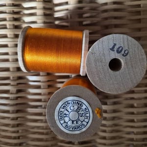 May include: Three wooden spools of thread. One spool is filled with bright orange silk thread. The top of the spool is stamped with the number 109 in black ink. Another spool has a label that reads "Rod Winding Silk" and "50 Yards".