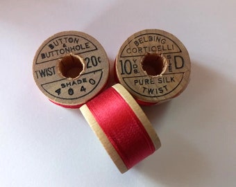Puede incluir: Tres carretes de hilo de madera vintage. Dos carretes redondos con texto impreso, uno dice "BUTTON & BUTTONHOLE TWIST" y el otro "BELDING CORTICELLI PURE SILK TWIST". También se ve un tercer carrete de hilo rojo.
