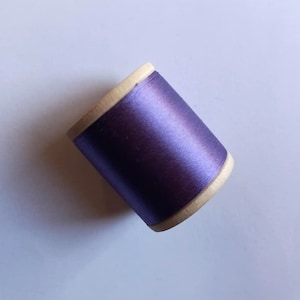 May include: A spool of dark purple thread with a subtle sheen. The thread is wound around a wooden core with a light tan color. The spool is set against a plain white background, highlighting the thread's color and texture.