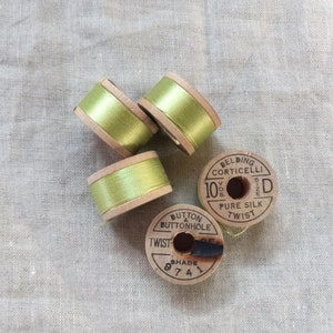 May include: Four spools of thread, three with bright green silk ribbon and wooden sides, and two vintage spools with text that reads "Belding Corticelli" and "Pure Silk Twist". The spools are on a light grey fabric.