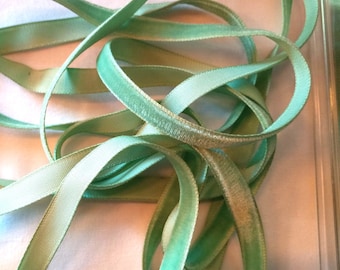 May include: A close-up of a light green satin ribbon with a velvet edge. The ribbon is twisted and folded, showing the texture of the fabric.