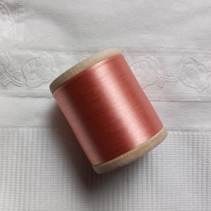 May include: A spool of coral-colored thread. The thread is tightly wound around a wooden core. The thread has a slight sheen, suggesting a silk or satin material. The spool is set against a white, textured background.