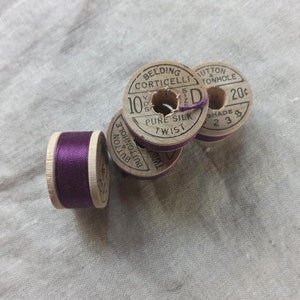 May include: A collection of vintage wooden spools of thread. The spools are labeled with text including "Belding Corticelli Pure Silk Twist". The thread is a deep purple color, and the spools are arranged on a light-colored fabric.