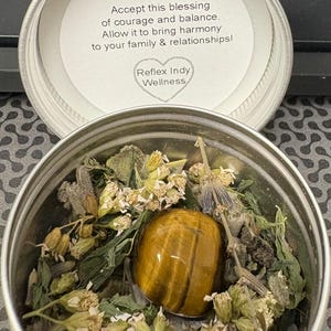 May include: A silver metal container with a lid. The lid has a white label with the text "Tigers Eye" and a message about courage and balance. The container is filled with dried herbs and a tiger's eye stone.