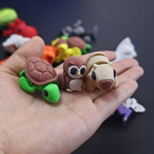 May include: A selection of small, colourful animal figurines. The foreground features a green turtle with a brown shell, a brown owl with large eyes, and a tan animal with black eyes. Other figurines are visible in the background.