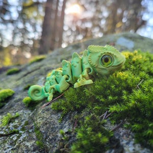 May include: A green chameleon figurine with a textured surface and large, yellow eyes. The figurine is posed on a bed of green moss.