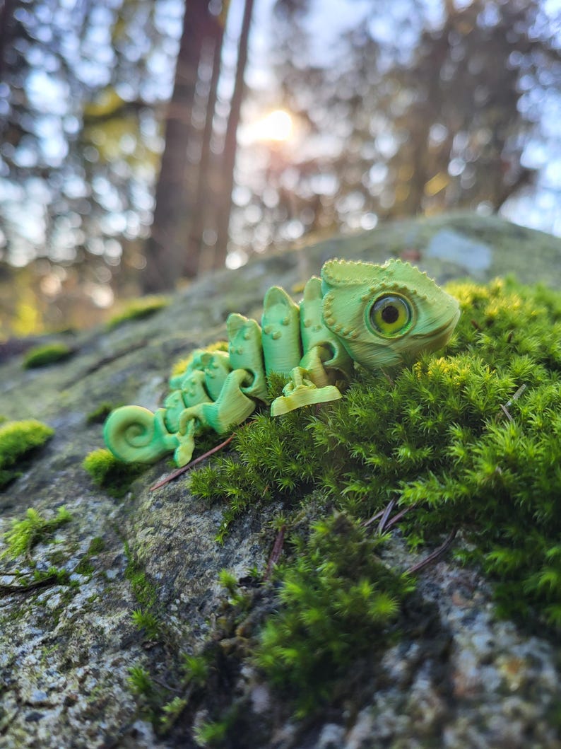May include: A green chameleon figurine with a textured surface and large, yellow eyes. The figurine is posed on a bed of green moss.