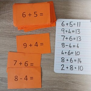 May include: Orange flash cards with addition and subtraction problems written in black marker. The cards are stacked on a wooden surface. A white board lists the answers to the problems on the cards. The text on the white board reads: "Printable Addition and Subtraction Flash Cards. Download and Print straight away as a PDF. 91 Addition facts. 55 subtraction facts. 146 facts in total. 8 cards per A4 page."