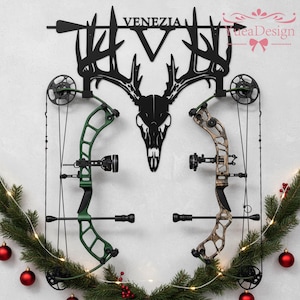 May include: A black metal wall mount featuring a deer skull and the word "VENEZIA". Two compound bows, one green and one camouflage, are mounted below. The display is decorated with a garland and red ornaments.