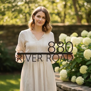 Peut inclure: Panneau d'adresse en métal noir avec les chiffres "880" au-dessus des mots "RIVER ROAD". Le panneau est tenu par une personne vêtue d'une robe crème, avec un arrière-plan vert flou.