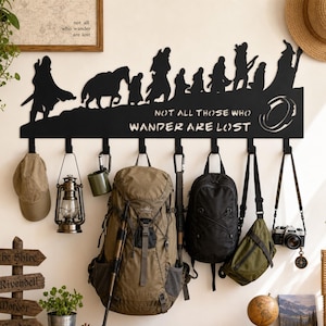 May include: A black metal wall-mounted coat rack featuring a silhouette of a group of travelers and the text "NOT ALL THOSE WHO WANDER ARE LOST" with a ring icon. The rack has eight hooks holding various items including a cap, lantern, and backpacks.