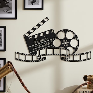 Könnte beinhalten: Schwarze Metall-Wandkunst mit einer Filmklappe, Filmrolle und Filmstreifen. Auf der Klappe steht "Luna's CINEMA". Filmrolle und Streifen sind dekorativ gestaltet. Ideal für Heimkino-Enthusiasten.
