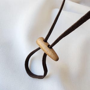 K&ouml;nnte beinhalten: Nahaufnahme eines Holzschalters mit zwei L&ouml;chern, befestigt an einer dunkelbraunen Kordel. Der Schalter ist hellbraun und zylindrisch, mit einer abgerundeten Form. Die Kordel ist geschlungen und scheint aus einem weichen, gewebten Material zu bestehen.