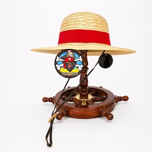 May include: A straw hat with a wide red ribbon band around the crown. A circular tag hangs from the brim featuring a cartoon monkey wearing a red bandana and goggles with the text MONKEY ANIME. A thin black chin strap with a wooden bead is attached.