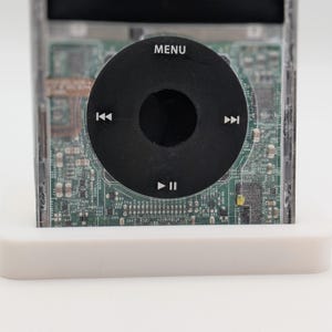 May include: A clear plastic case with a green circuit board inside. The case has a black circular button with the words "MENU", "<<", ">>", and "Play/Pause" printed on it.
