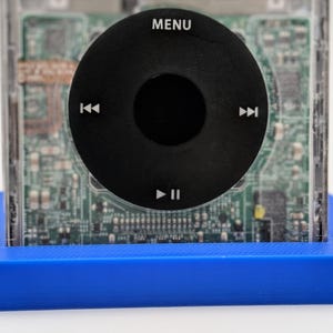 May include: A blue plastic base with a clear plastic case on top. The case contains a circuit board with a black circular button with the words "MENU", "<", "||", ">" printed on it.
