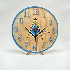 May include: A wooden clock with a blue border and Roman numeral hour markers. The clock face features a yellow and blue Masonic symbol.